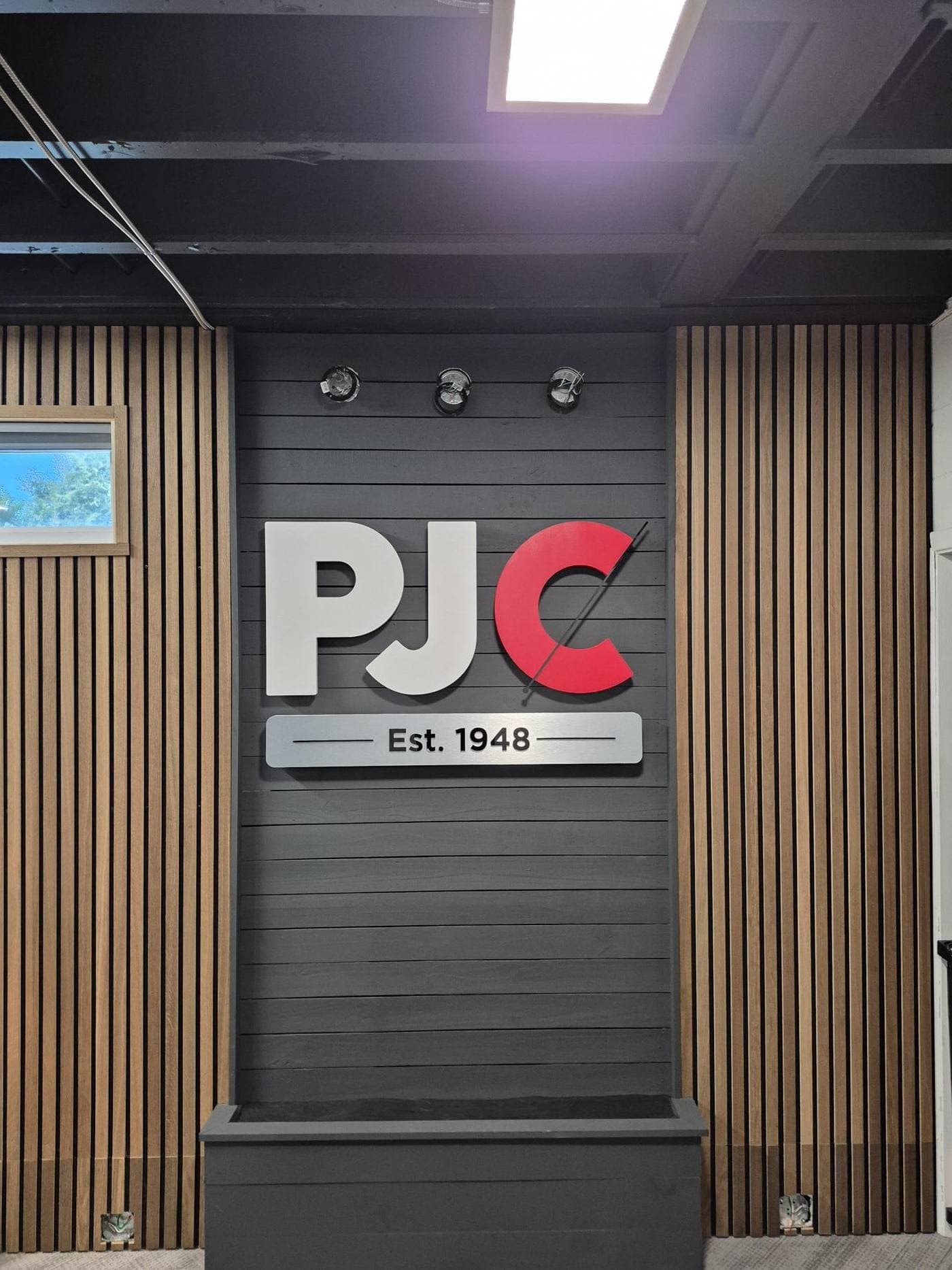 Oversized painted white and red dimensional letters with a brushed metal-finish founding-date plaque on a dark wood-slat feature wall in a Tampa office lobby.